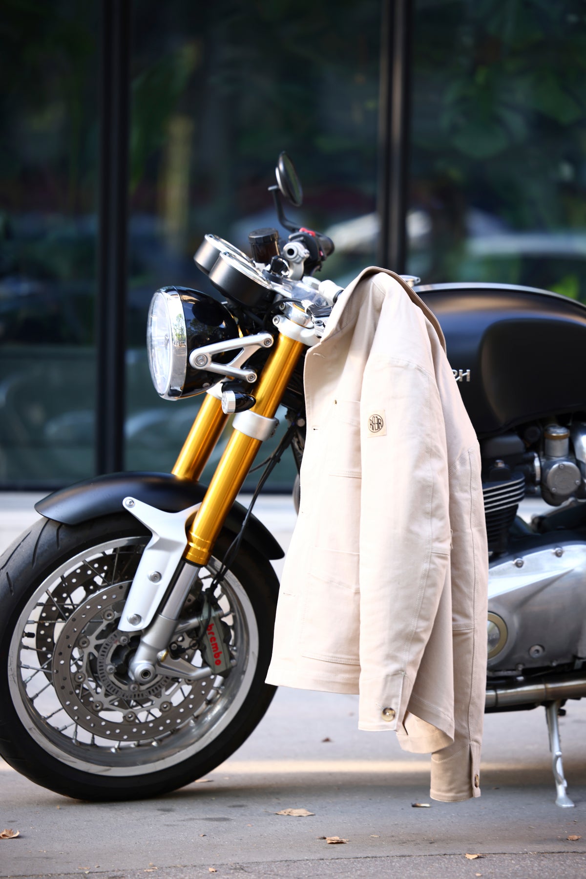 Veste ARTIST beige posée sur une moto, détails du tissu et écusson RAER