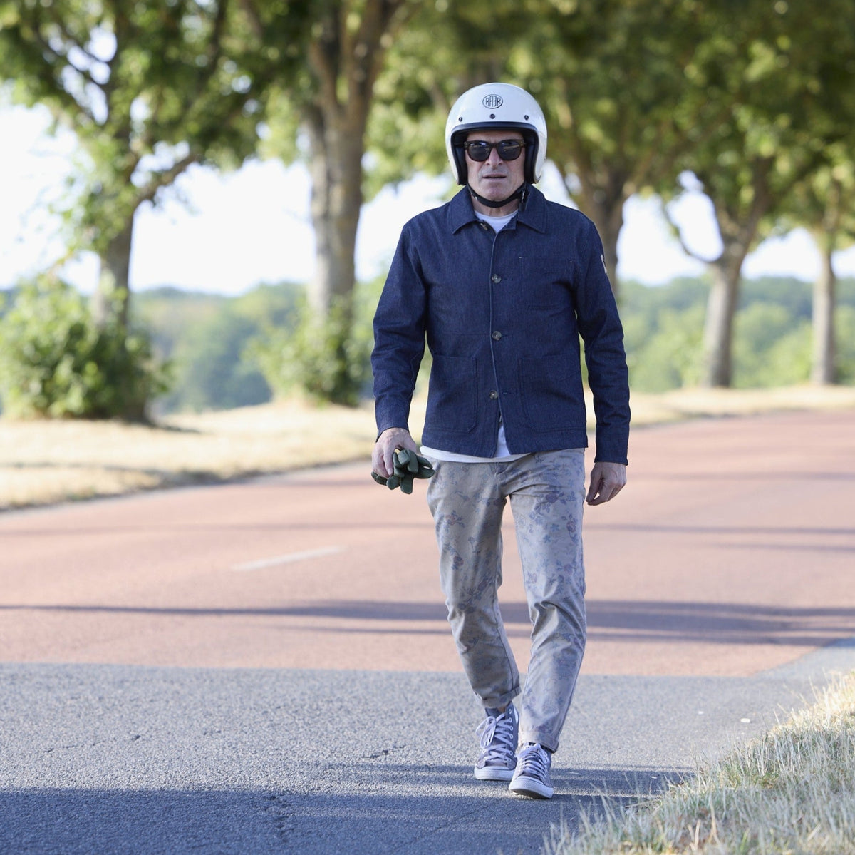 Homme portant une veste RAER bleu indigo, casque blanc et gants à la main sur une route bordée d’arbres