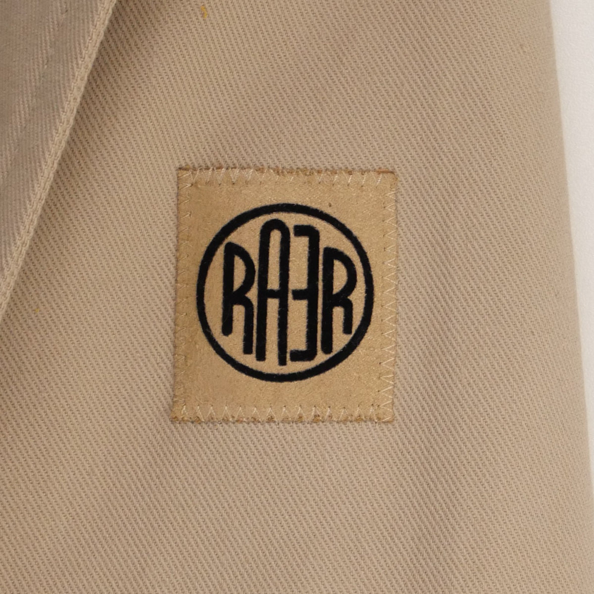 Gros plan du logo RAER cousu sur la toile beige de la veste ARTIST.