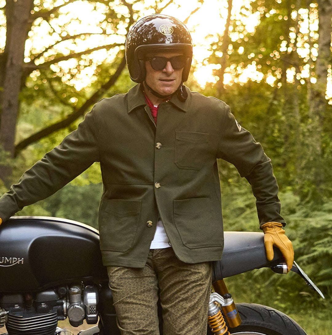 Veste ARTIST kaki portée de face par un homme casqué à côté d’une moto.