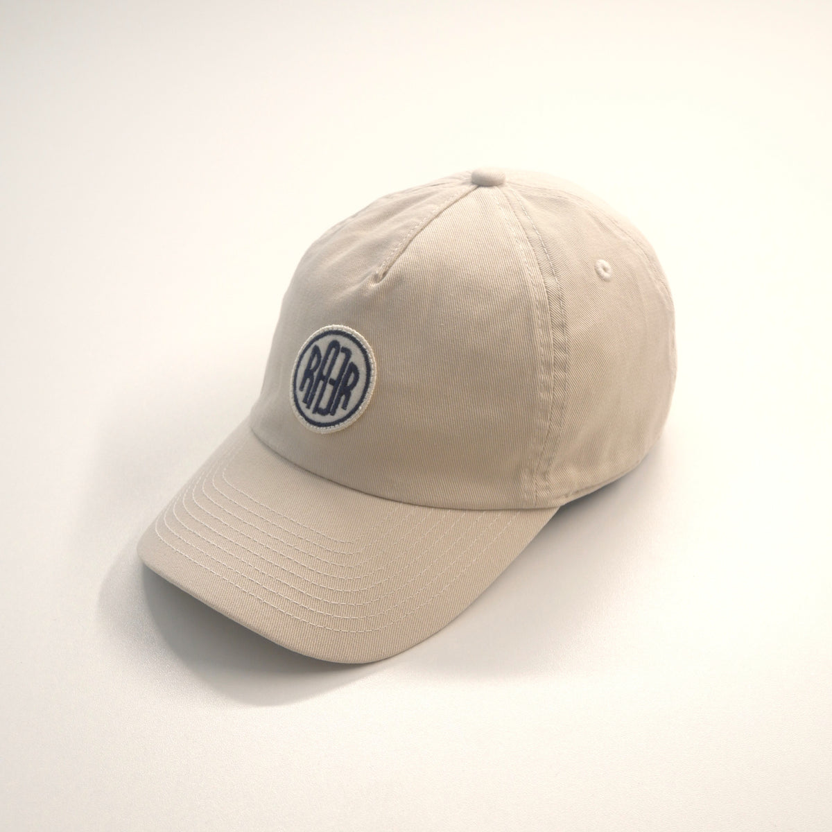 Casquette ORIGINAL écrue vue de trois quarts, avec badge RAER à l’avant et visière courbe.