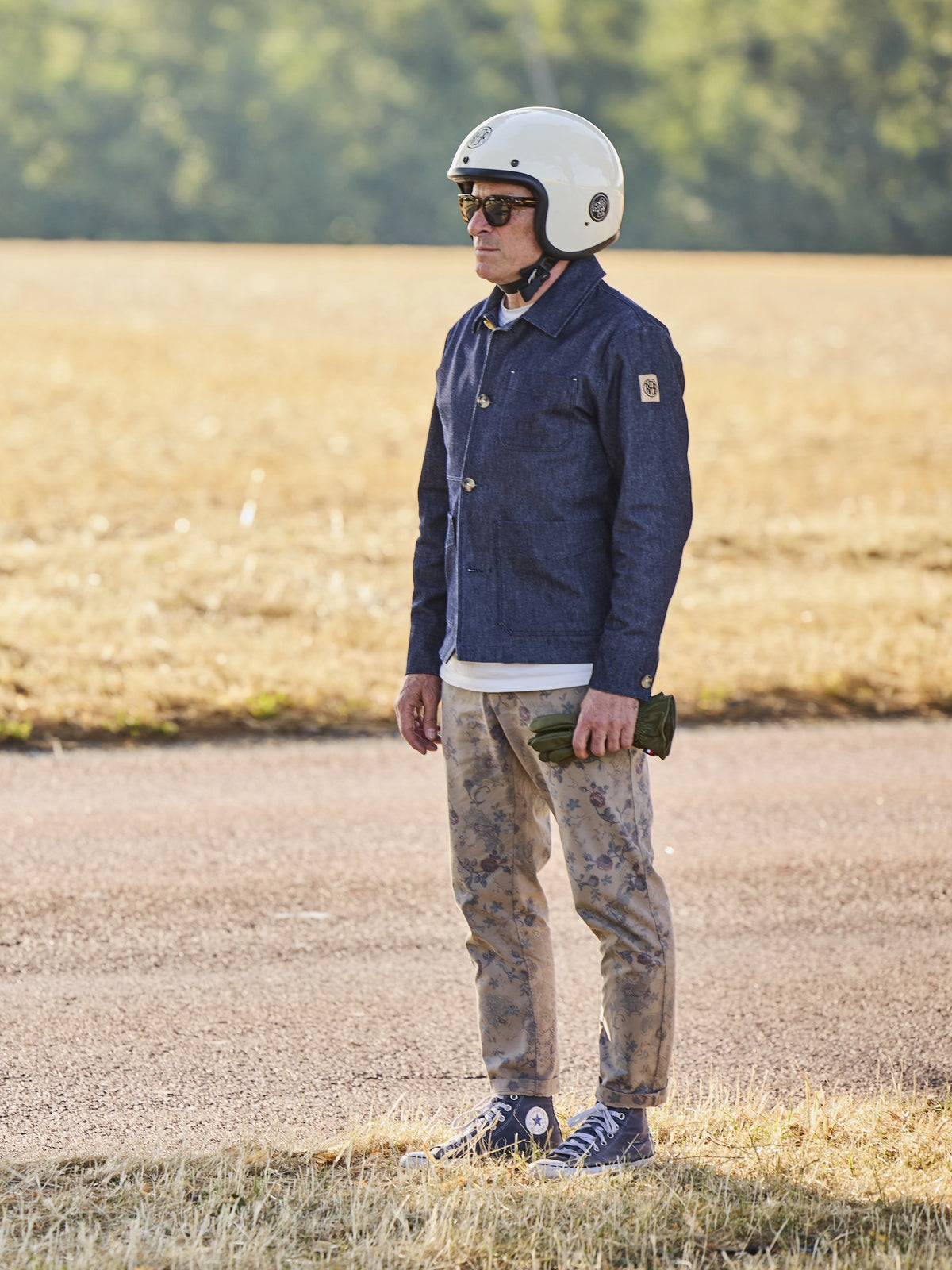 Homme portant une veste RAER bleu indigo, casque blanc et gants à la main sur une route bordée d’arbres