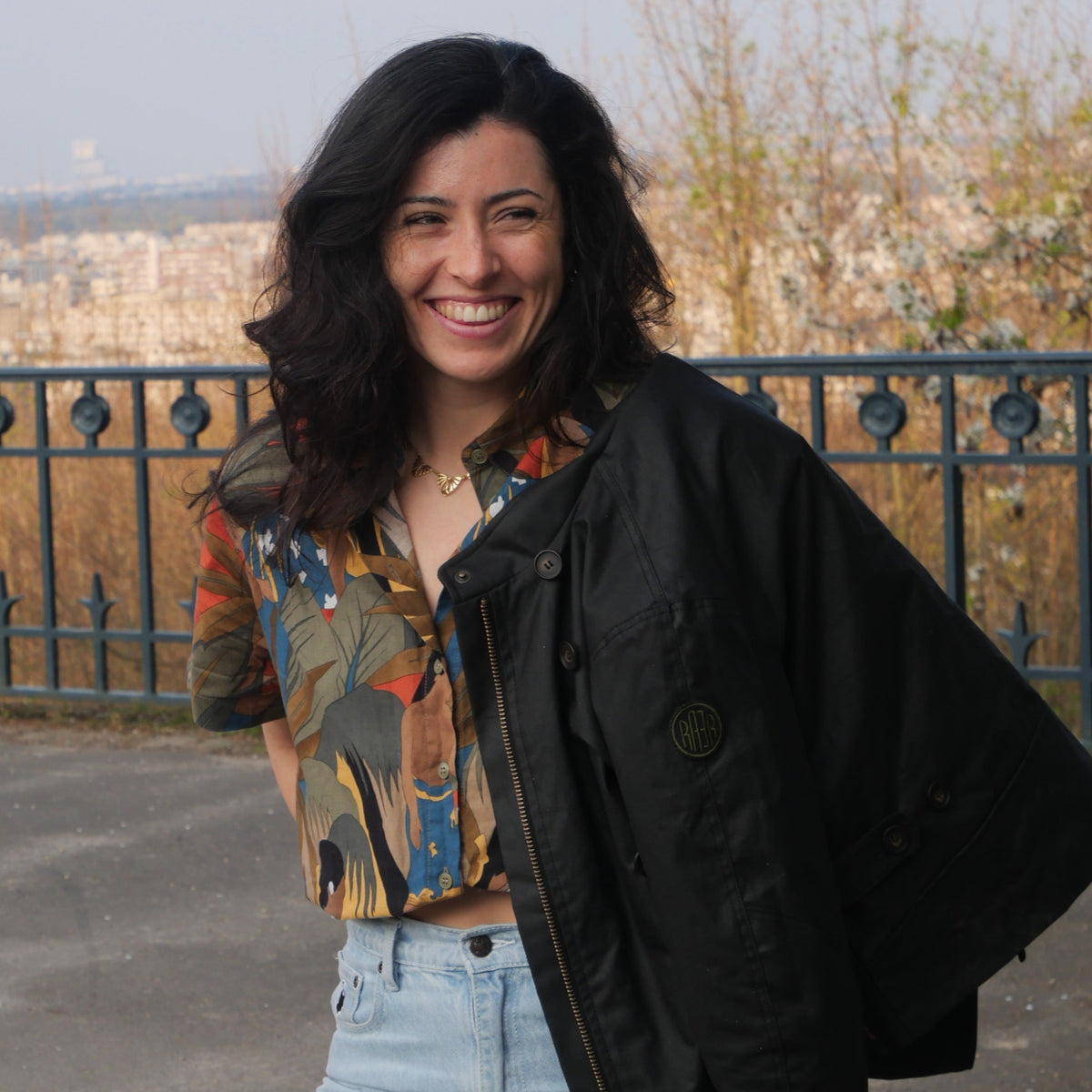 Veste DONNA Wax noire portée ouverte sur une blouse imprimée, en extérieur.